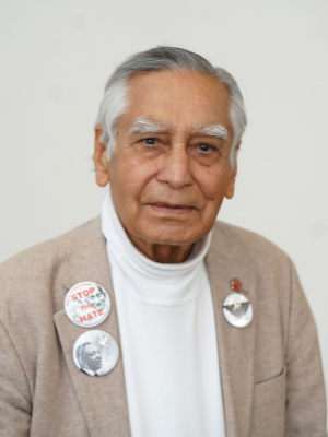 Dr. Sudarshan Kapoor wears a tan sports jacket over a white turtleneck shirt. The jacket has three peace buttons, "Stop the Hate," a Dr. Martin Luther King, Jr. button and a United Farm Workers Button.