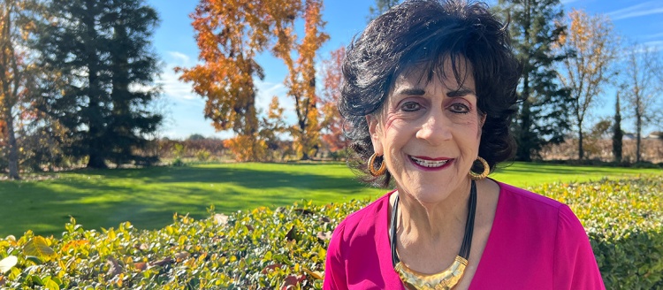 Jane Bedrosian in a pink top and standing in front of a lawn with fall leave trees in the background.