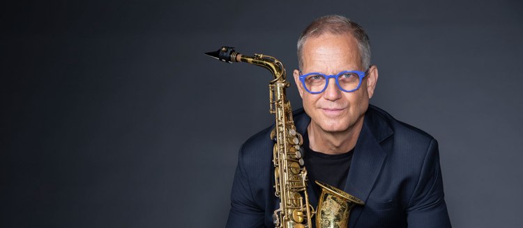 Benjamin Boone holds his saxophone in front of a dark grey-blue background wearing a dark blue sport jacket and black Tshirt with bright blue eyeglasses.