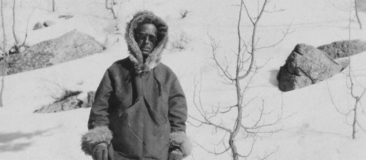 Black and white photo of Orland Bartholomew in the mountains wearing a large fir-lined coat.
