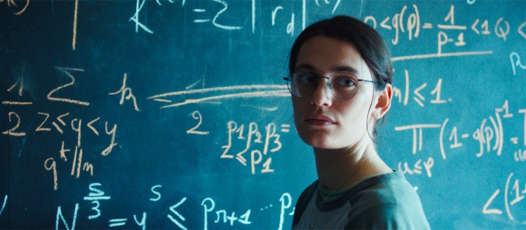 A women with dark hair and glasses stands in front of a chalk board with many mathematical equations on it. She is wearing a t-shirt and half of her face is illuminated while half is in the dark.