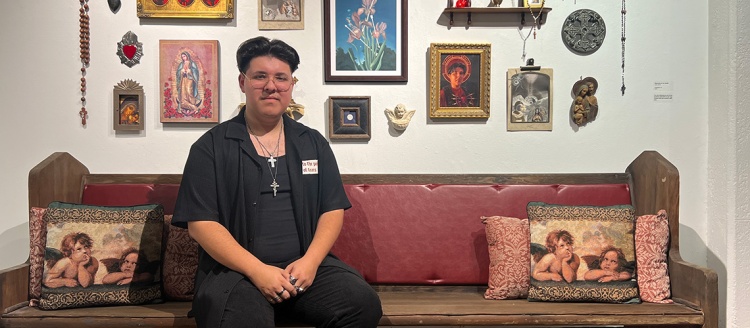 Jose Soria in black with two cross necklaces sits on a pew in front of his Catholic religious themed artwork. with at his exhibition, "I Exist Both Here and There"