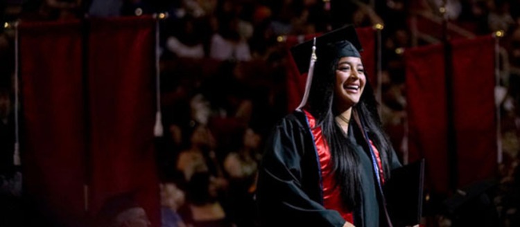 A Fresno State Graduate in a black cap and gown with a red and blue sash is illuminated by lights during graduation.